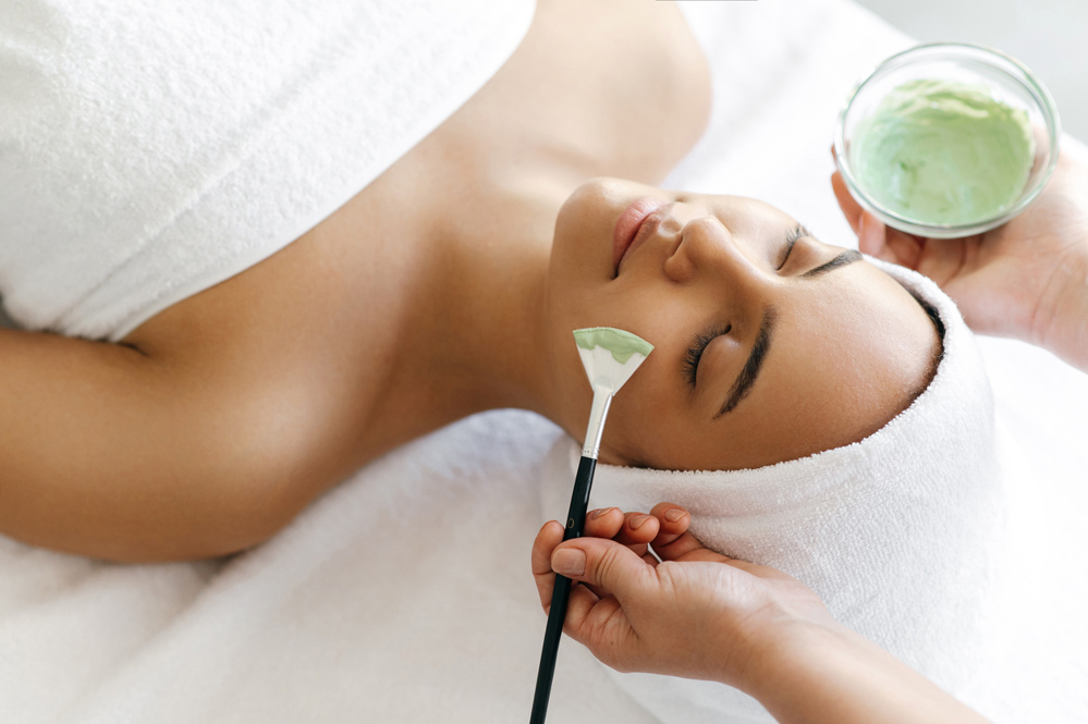 Facial treatments, wrinkle prevention, facial cleansing. Top view of beauty procedure, therapist applying green face mask on the face of a beautiful brazilian or hispanic young woman
