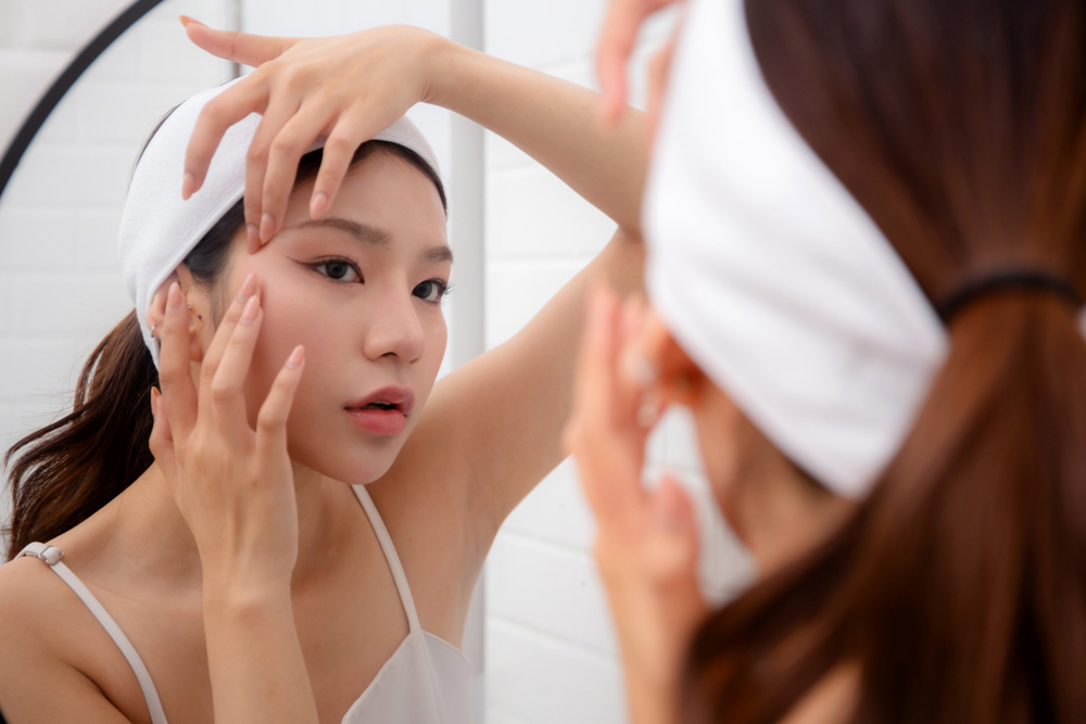Beautiful young asian woman examining skin of facial while looking mirror in bathroom at home, unhappy woman touching cheek with check wrinkle with stress and worry, beauty and skincare concept
