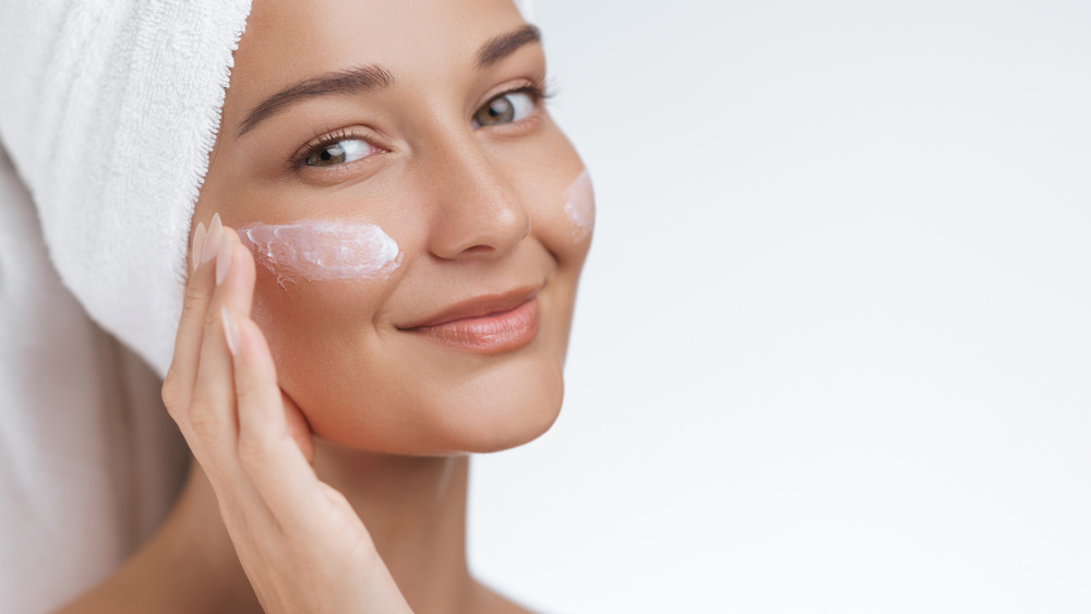 Close-up of a smiling woman with a towel wrapped around her hair, applying face cream as part of her skincare routine. Ideal for showcasing beauty, wellness, and self-care practices focused on maintaining healthy, radiant skin